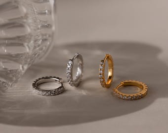 May include: A collection of four small hoop earrings, two in a silver tone and two in a gold tone. Each hoop features a row of small, clear, sparkling stones embedded along the front-facing surface. The earrings are arranged in a cluster.