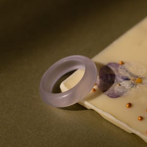 Può includere: Un anello lilla chiaro dal design liscio e arrotondato. L'anello è appoggiato su una superficie bianca con un fiore viola pressato e piccole decorazioni color oro. Lo sfondo è verde tenue.