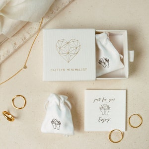 May include: A white box with a gold geometric heart design and the text "CAITLYN MINIMALIST". The box is open and contains a small white pouch with a gold heart design and the text "just for you! Enjoy!" on a separate card.