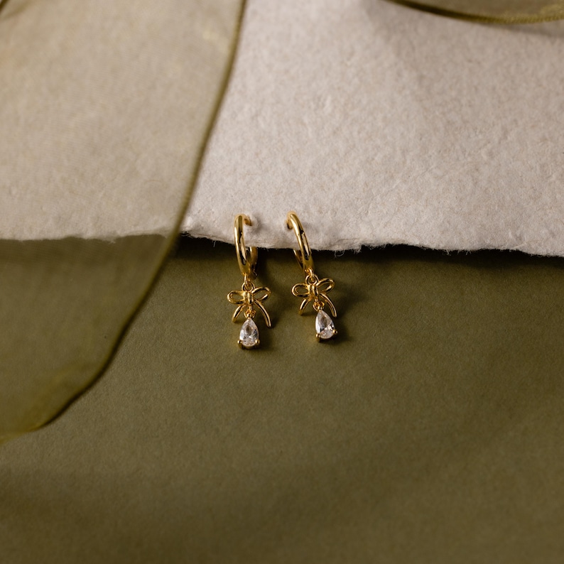 May include: A pair of gold-colored hoop earrings. Each earring features a bow-shaped charm and a teardrop-shaped, clear gemstone pendant. The earrings are displayed on a textured, off-white surface against a muted green background.