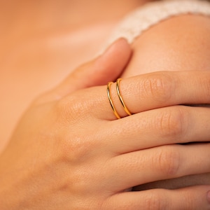 May include: Close-up of two thin, gold-colored rings worn on a finger. The rings are simple bands, reflecting light. The image focuses on the jewelry, highlighting the minimalist design and the warm, golden hue of the metal.