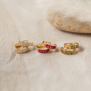 May include: Three pairs of gold hoop earrings, each featuring a row of small square-cut stones. One pair has clear white stones, one pair has vibrant pink stones, and the third pair has deep olive green stones, all set in a polished gold metal finish.