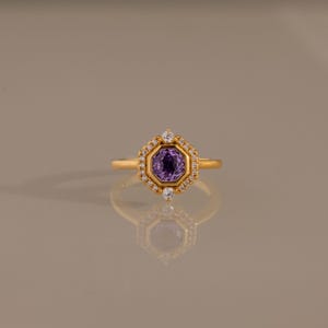 May include: A gold ring featuring a central octagonal purple gemstone surrounded by a halo of small clear stones. The setting is hexagonal in shape with two larger clear stones positioned at the top and bottom of the central purple gem.