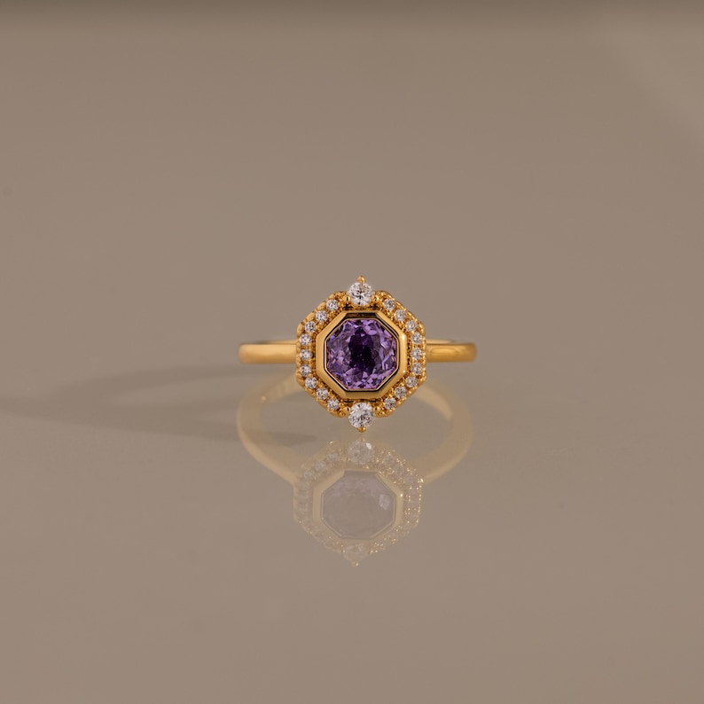 May include: A gold ring featuring a central octagonal purple gemstone surrounded by a halo of small clear stones. The setting is hexagonal in shape with two larger clear stones positioned at the top and bottom of the central purple gem.