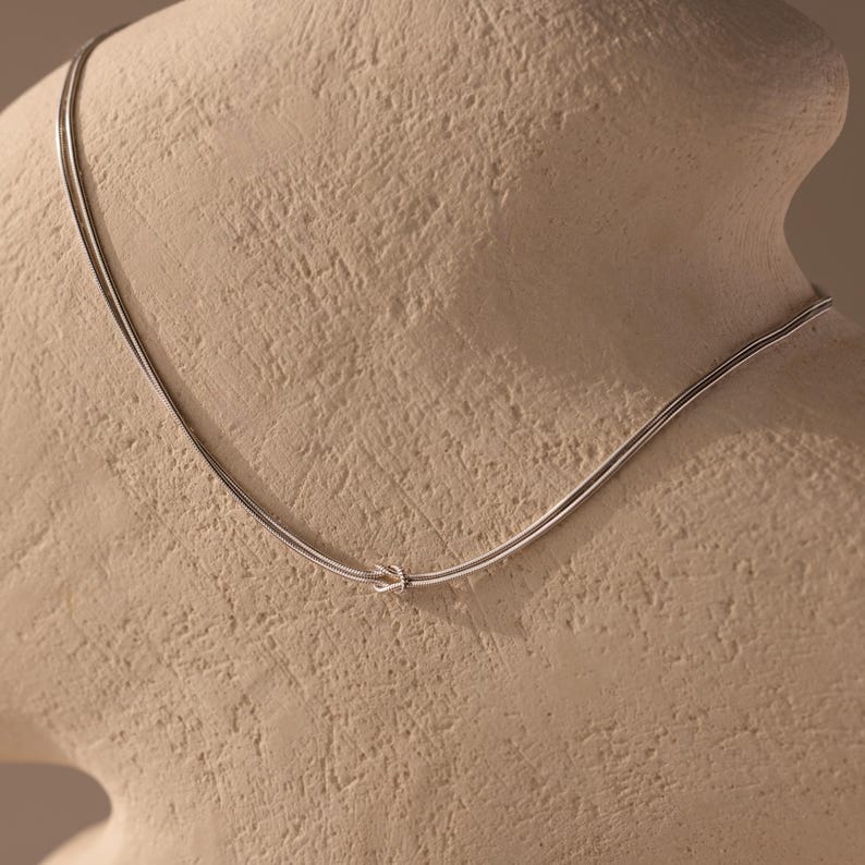 May include: A delicate silver necklace with a thin, double-strand design. The necklace features a small, circular clasp at the center, and is displayed on a neutral-toned mannequin. The jewelry is simple and elegant.