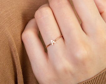 May include: A delicate rose gold ring featuring the letter "A" in a minimalist style. The ring is thin and elegant, perfect for everyday wear. The ring is shown on a hand with a neutral-toned background.