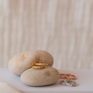 May include: Gold hoop earrings with small, clear gemstones are displayed on a light-colored stone. Additional earrings in rose gold and silver are visible in the background. The earrings are a jewelry item.