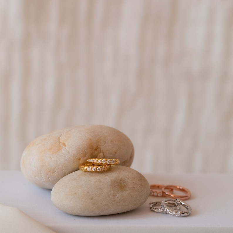 May include: Gold hoop earrings with small, clear gemstones are displayed on a light-colored stone. Additional earrings in rose gold and silver are visible in the background. The earrings are a jewelry item.
