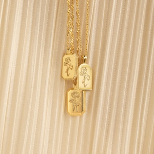 May include: Three gold-toned necklaces featuring pendant charms with engraved floral designs. Two pendants are rectangular with textured edges, and one is an arched shape. Each pendant displays a different botanical illustration on a polished surface.