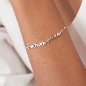 May include: A silver bracelet with the names "Hudson" and "Alexa" in a delicate script font. The bracelet is shown on a person's arm, with a white background.