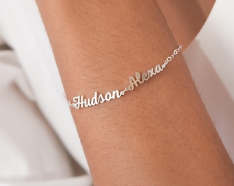 May include: A silver bracelet with the names "Hudson" and "Alexa" in a delicate script font. The bracelet is shown on a person's arm, with a white background.