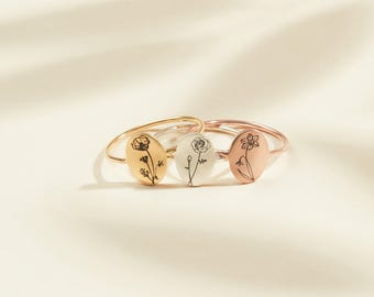 Pode incluir: Três anéis finos de metal com pingentes ovais. Cada pingente é gravado com um design floral distinto em linhas pretas. Os anéis têm acabamentos em tons de ouro, prata e ouro rosa, dispostos em um grupo para exibir as gravuras botânicas.