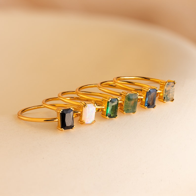 May include: Six gold rings with rectangular gemstones in various colors: black, white, green, teal, blue, and a mossy green. The rings are arranged in a row, showcasing the different gemstone options.