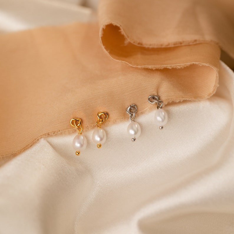 May include: Close-up of four earrings with a pearl drop. Two earrings have gold-colored heart-shaped tops, and two have silver-colored tops. The earrings are displayed on a white and tan fabric background.