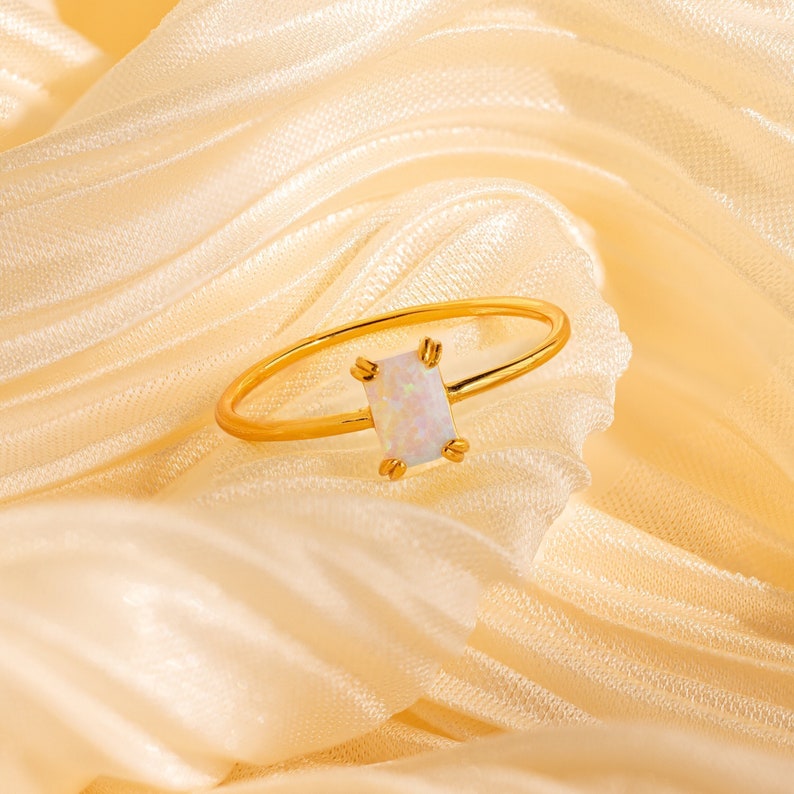 May include: A gold-colored ring with a rectangular, iridescent opal stone. The ring is set on a soft, cream-colored fabric, highlighting the ring's delicate design. The opal stone is the focal point, showcasing its subtle color variations.