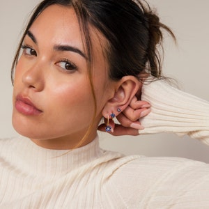 May include: A woman wearing a white sweater with blue flower earrings. The earrings are gold and have small blue flowers with white centers.