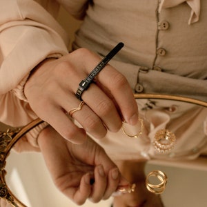 May include: A close-up of a hand wearing a ring sizer tool and several gold rings. The hand is resting on a mirrored surface with other gold rings reflected in the mirror.