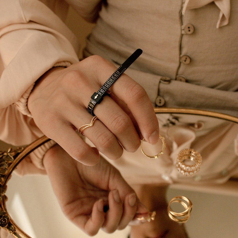May include: A close-up of a hand wearing a ring sizer tool and several gold rings. The hand is resting on a mirrored surface with other gold rings reflected in the mirror.