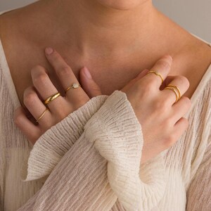 May include: A person's hands wearing multiple gold rings. The rings are thin and delicate, with some featuring small stones. The person is wearing a white sweater.