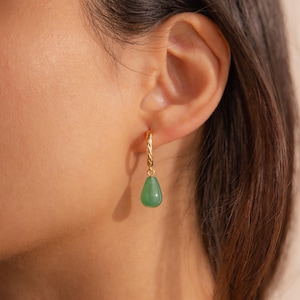 May include: A gold hoop earring with a twisted texture featuring a single smooth, teardrop-shaped green stone pendant hanging from the bottom. The earring is secured through the earlobe, showcasing a polished metallic finish and a vibrant green hue.