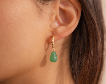 Può includere: Un orecchino a cerchio dorato con una texture intrecciata, caratterizzato da un singolo ciondolo in pietra verde liscia a forma di goccia che pende dal fondo. L'orecchino è fissato al lobo, mostrando una finitura metallica lucida e una tonalità verde vivace.