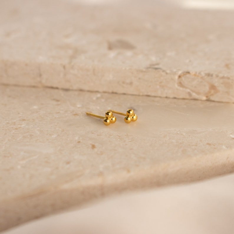 May include: A pair of gold-toned stud earrings with three small, round beads clustered together.