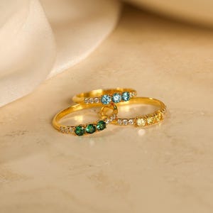May include: Three gold rings, each featuring a trio of round-cut gemstones in green, blue, and yellow, accented by small clear stones along the band. The rings are arranged in a cluster, showcasing the polished metal finish and the vibrant colors of the stones.