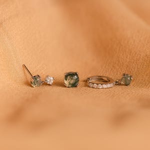 May include: Three silver earrings with green moss agate stones and clear cubic zirconia accents. The earrings are on a light brown surface.