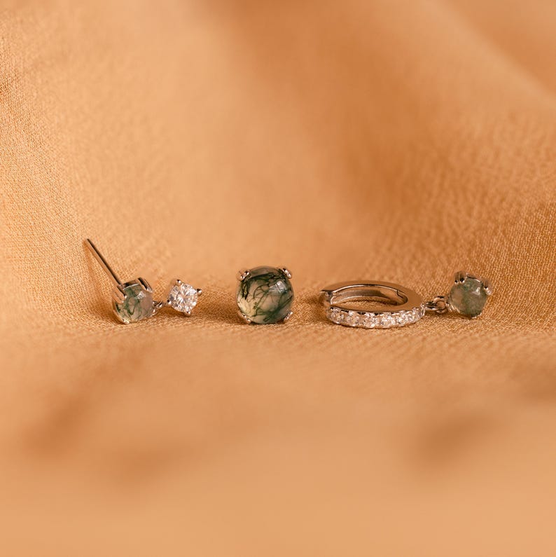 May include: Three silver earrings with green moss agate stones and clear cubic zirconia accents. The earrings are on a light brown surface.