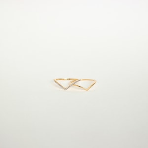 Può includere: Due anelli dorati a forma di V. Un anello è adornato con piccole pietre preziose trasparenti lungo la forma a V, mentre l'altro è in oro lucido semplice. Entrambi gli anelli sono su uno sfondo bianco.