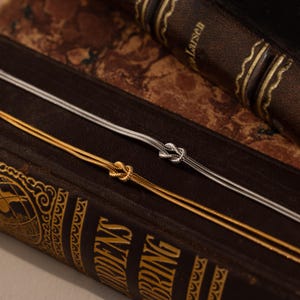 May include: Two delicate necklaces, one gold and one silver, each featuring a knot design. The necklaces are displayed on a vintage book with gold lettering and a dark brown leather cover. The gold necklace is on the bottom, and the silver necklace is on top.