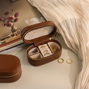 May include: A brown leather jewelry box with a zipper closure. The box is open and has multiple compartments for rings, earrings, and other jewelry. The box has the words 'Carrie Elizabeth' embossed on the top.