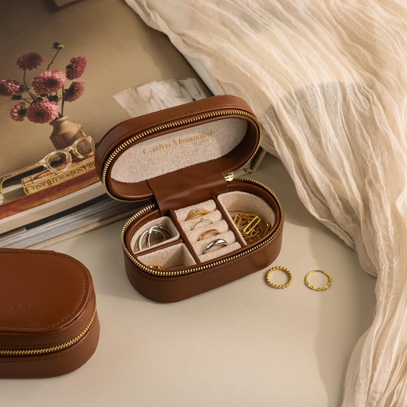 May include: A brown leather jewelry box with a zipper closure. The box is open and has multiple compartments for rings, earrings, and other jewelry. The box has the words 'Carrie Elizabeth' embossed on the top.