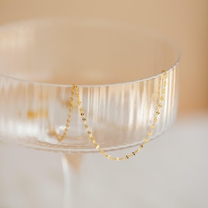 May include: A delicate gold necklace with a chain of small, flat, circular links hangs over the rim of a clear glass. The necklace is suspended in front of a soft, blurred background, creating a minimalist aesthetic.