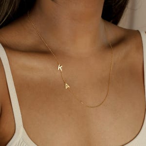 Puede incluir: Un delicado collar dorado con las letras "K" y "A". El colgante está unido a una fina cadena, creando un diseño minimalista y elegante. El collar se lleva alrededor del cuello.