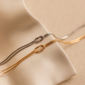 May include: Two delicate gold and silver chain necklaces with a knot design. The necklaces are displayed on a beige fabric background.