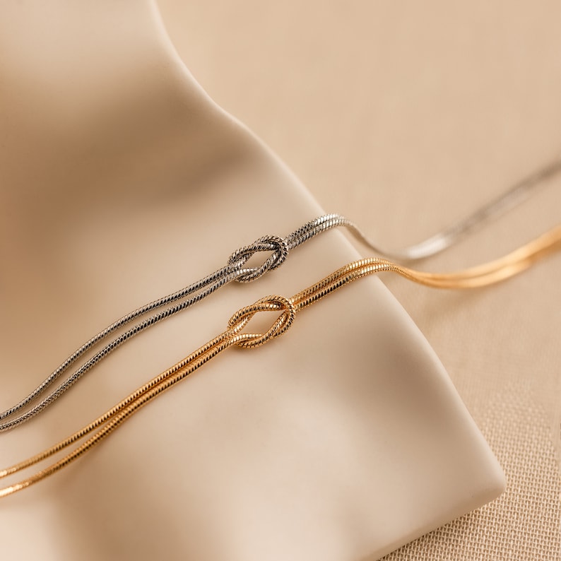 May include: Two delicate gold and silver chain necklaces with a knot design. The necklaces are displayed on a beige fabric background.