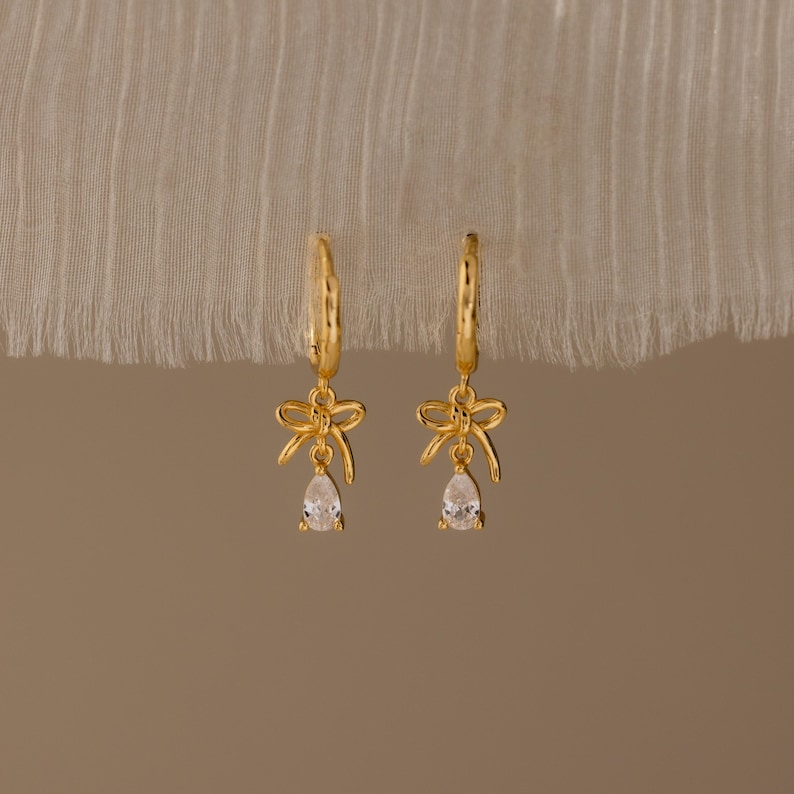 May include: A pair of gold-colored hoop earrings with bow-shaped charms and teardrop-shaped clear gemstones. The earrings are displayed against a neutral-toned background, highlighting their delicate design and sparkle.