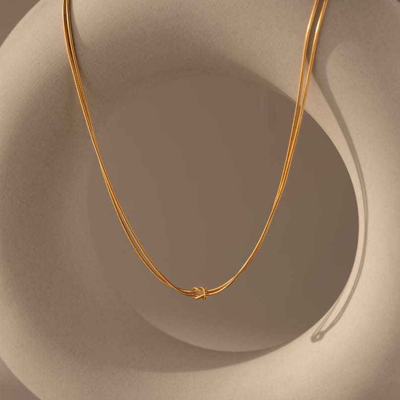May include: A delicate gold necklace with a knotted design. The necklace features two fine strands of gold that meet at a small knot in the center. The jewelry is displayed against a neutral background, highlighting its elegant simplicity.