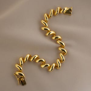 May include: A gold bracelet with a distinctive, undulating design. The bracelet comprises a series of connected, curved links, forming a flowing, attractive pattern. The clasp is rectangular and gold-toned. The jewellery item is presented against a neutral, beige backdrop.