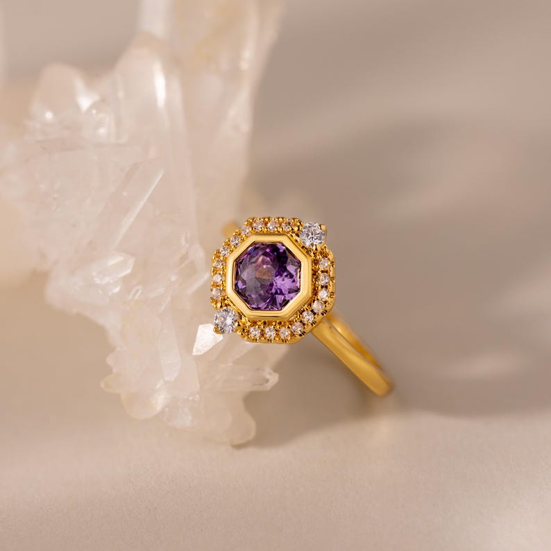 May include: A gold ring featuring an octagonal purple gemstone at the centre, surrounded by a halo of small clear stones. The band is smooth and polished gold, and the setting is designed with a textured, beaded edge around the central stone.