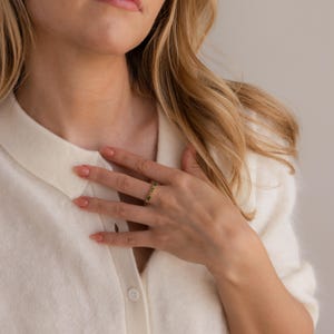 May include: A gold ring with small, dark green gemstones set around the band. The ring is on a person's finger, with the hand resting on a white sweater. The ring is a piece of jewelry.