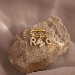 Op de afbeelding: Een goudkleurige, ovale karabijnsluiting met drie bungelende letterbedels, R, W en O, elk ingelegd met kleine doorzichtige steentjes. De metalen bedels hebben een pavé-afwerking en hangen aan de onderkant van de ovale ring.