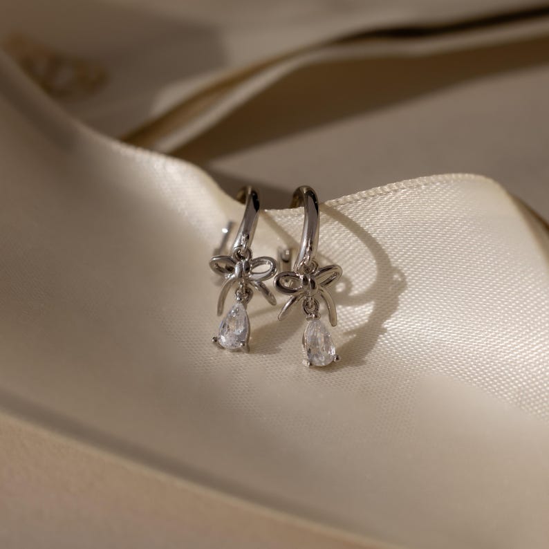 May include: A pair of silver hoop earrings with a bow design and a teardrop-shaped clear gemstone. The earrings are displayed on a cream-colored satin fabric.