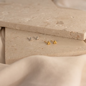 May include: Two pairs of small, simple stud earrings. One pair is silver and the other is gold. Each earring has three small, round beads.