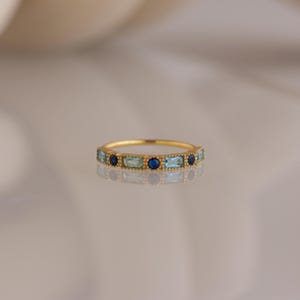 May include: A gold ring featuring alternating blue and light blue gemstones. The ring has a delicate design with rectangular and round stones set in a gold band. The gemstones are evenly spaced around the ring.