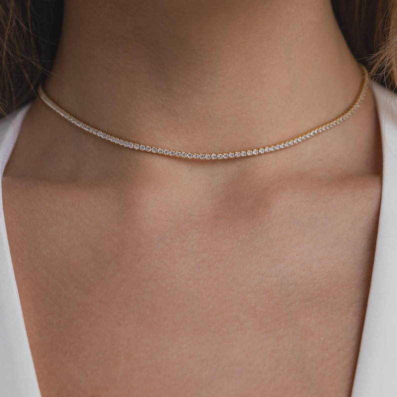 May include: A delicate gold necklace with a row of small, sparkling white gemstones. The necklace is worn around the neck, resting on the collarbone. The background is a soft, neutral color, highlighting the jewelry.