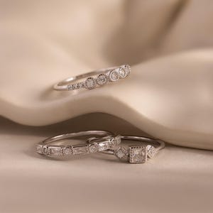 May include: Three silver rings with clear gemstones. The top ring has a series of round stones. The middle ring has a mix of rectangular and round stones. The bottom ring features a square stone flanked by smaller square and diamond-shaped stones.