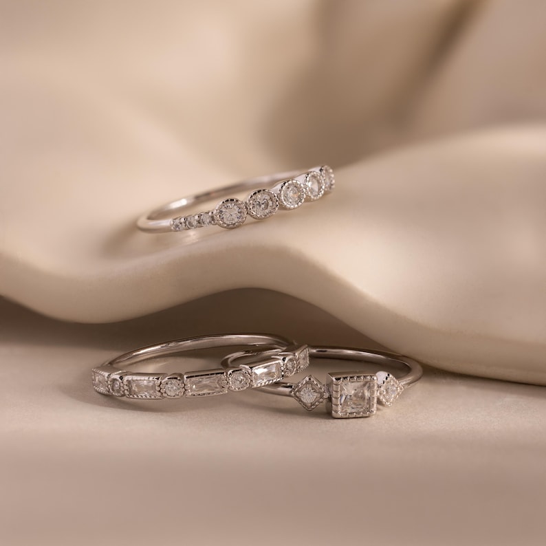 May include: Three silver rings with clear gemstones. The top ring has a series of round stones. The middle ring has a mix of rectangular and round stones. The bottom ring features a square stone flanked by smaller square and diamond-shaped stones.