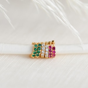 May include: A collection of six small gold hoop earrings arranged in a row. Two hoops feature green stones, two feature clear white stones, and two feature vibrant pink stones. Each hoop is circular with a hinged closure and a row of small gems.
